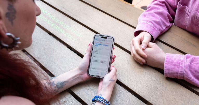 Close up of two people's hands resting on table opposite each other, one has tattoos on arms and is holding a phone with the Talk to Us page open