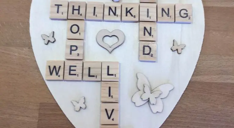 A heart-shaped white plaque features wooden Scrabble tiles forming the words "THINKING," "WELL," "KIND," and "LIVE." The word "THINKING" is horizontally placed at the top, intersecting with "KIND" vertically. "WELL" is arranged horizontally at the bottom, while "LIVE" is positioned vertically, intersecting with "WELL." In the center of the plaque is a cut-out heart shape, and surrounding the words are small, wooden butterfly decorations. The plaque is set against a light wooden surface.