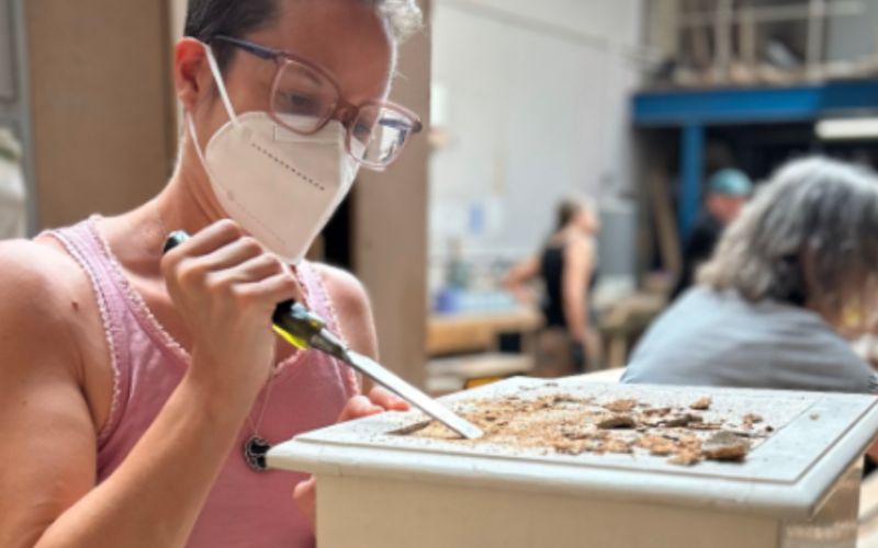 A person in a pink tank top and glasses is wearing a face mask while working on a furniture restoration project. They are using a chisel to scrape material from the surface of a wooden object. In the background, other people are also engaged in similar restoration activities in a workshop setting.