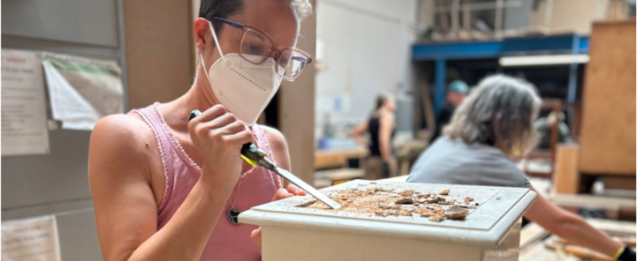 A person in a pink tank top and glasses is wearing a face mask while working on a furniture restoration project. They are using a chisel to scrape material from the surface of a wooden object. In the background, other people are also engaged in similar restoration activities in a workshop setting.