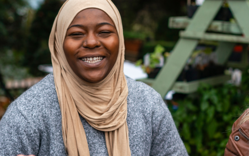 Three women - one in a beige headscarf, one with a burnt orange cap and one with her side profile visible, engaged in conversation, sharing thoughts and smiles in a vibrant setting.