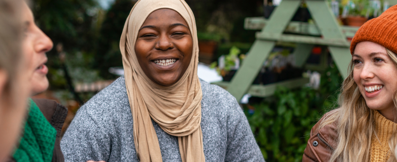 Three women - one in a beige headscarf, one with a burnt orange cap and one with her side profile visible, engaged in conversation, sharing thoughts and smiles in a vibrant setting.