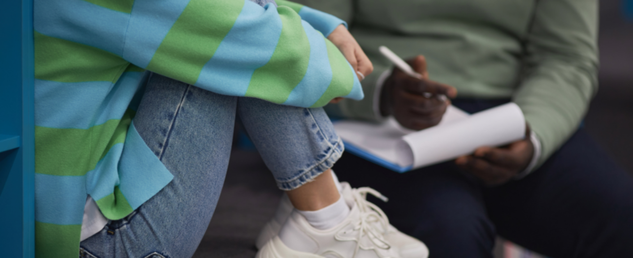 An indicative image of two people - one taking notes on a notepad while the other sits with their hands wrapped around their knees. the image is cropped to not see the faces.